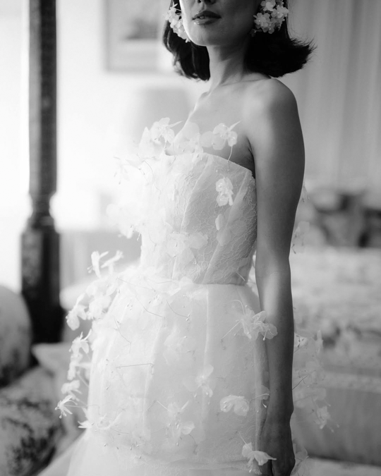 A black and white photograph shows a person in an elegant white wedding dress with delicate ruffled details.
