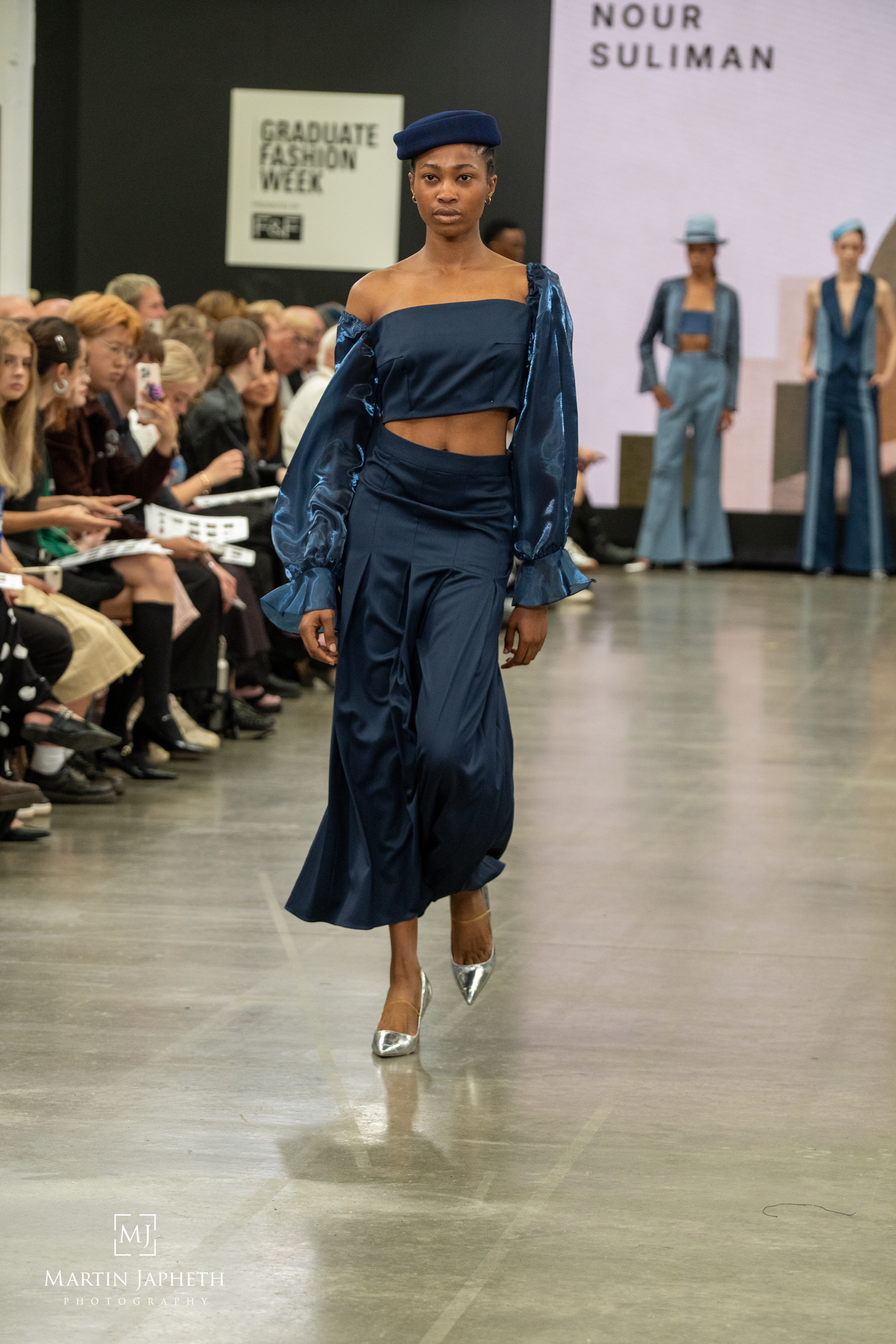 Model walks runway in navy blue satin crop top, wide-leg pants, and beret at Graduate Fashion Week show. Martin Japheth Photography