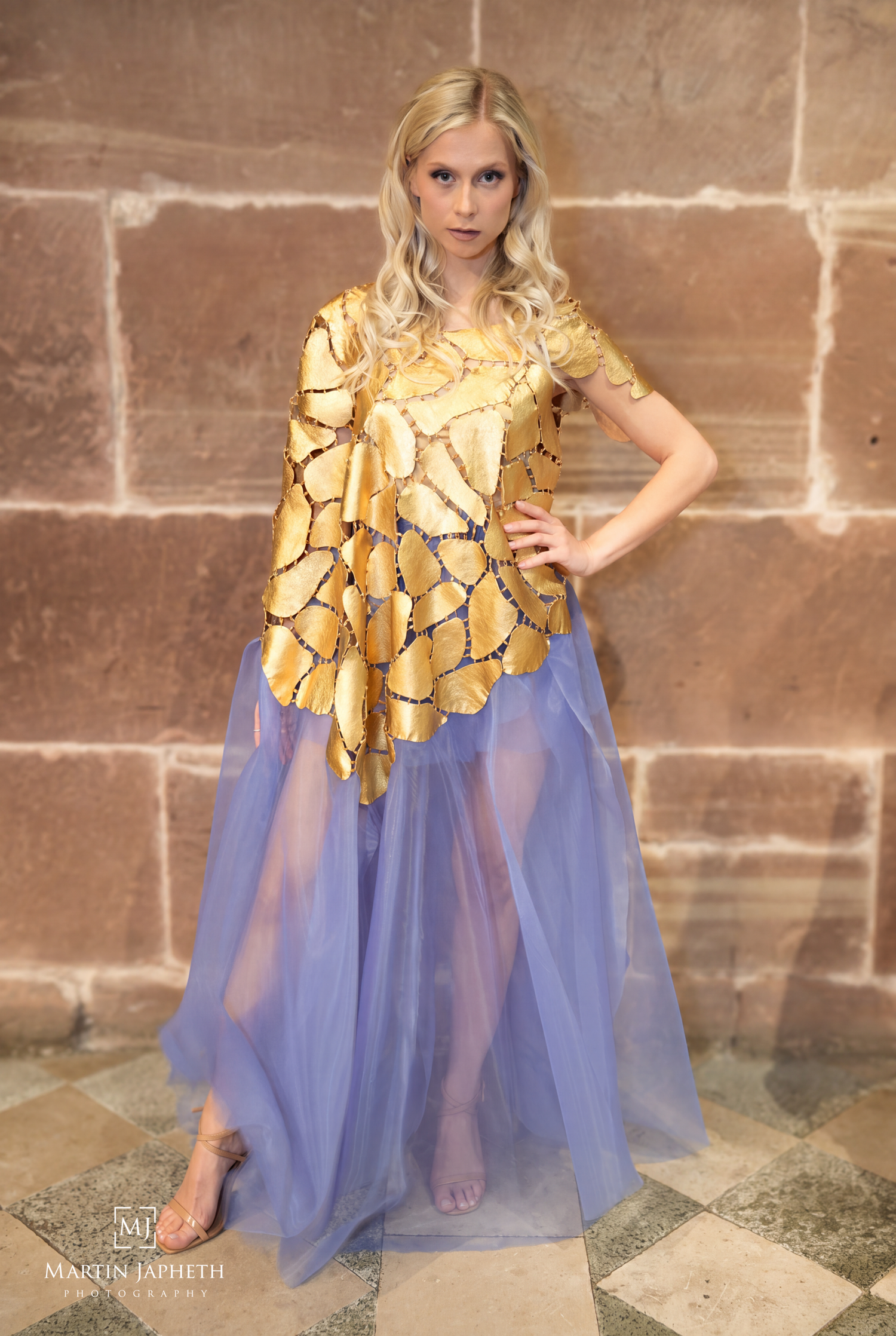 Blonde woman in gold sequined top and sheer lavender skirt posing against stone wall.
