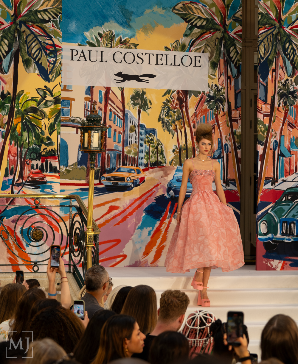Models walk the runway showcasing colorful evening gowns at a fashion show with palm tree backdrop decor.