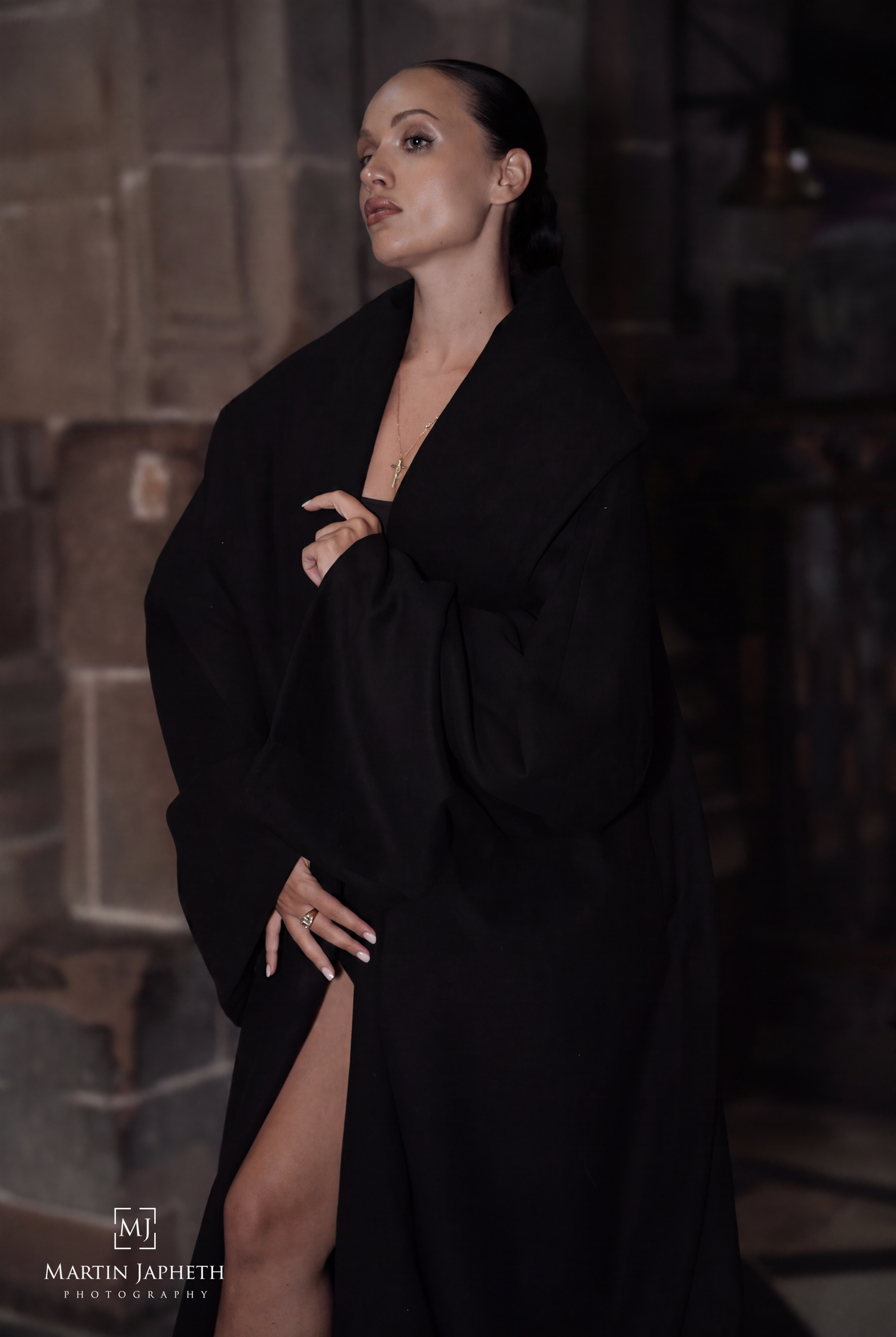 Woman in elegant black robe poses dramatically against stone wall in moody, artistic fashion photography.