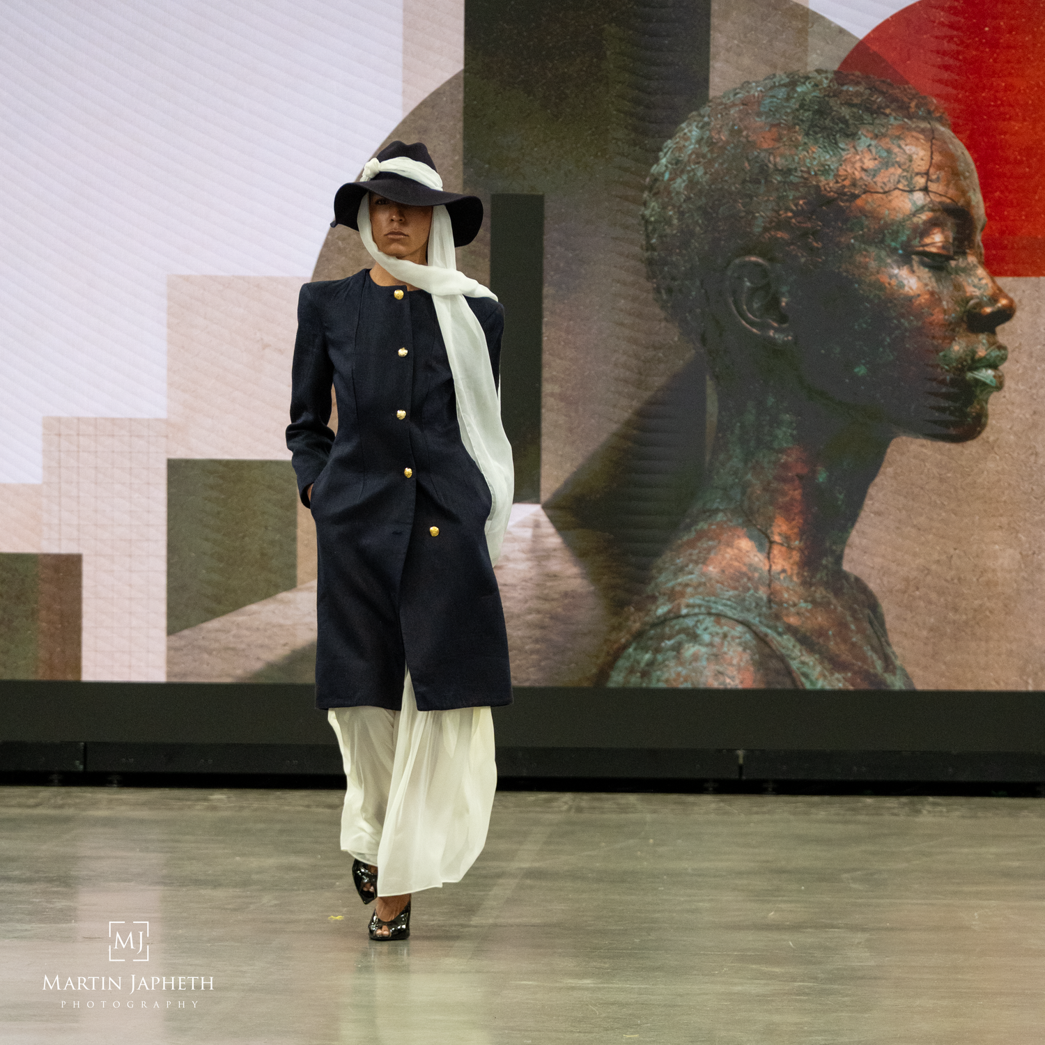Model walks runway in navy coat, white wide-leg pants, and white hat before a large portrait Martin Japheth Photography