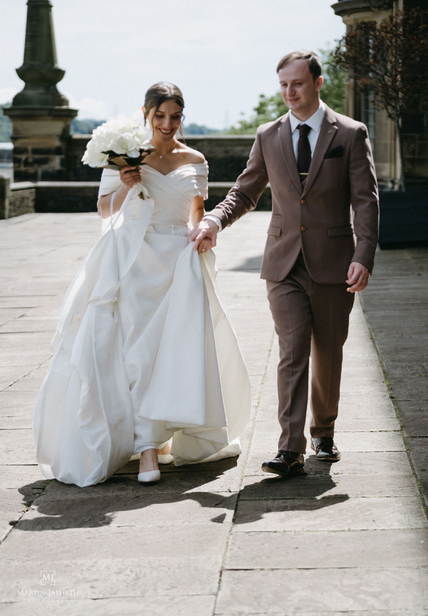 Bride in off-shoulder white gown holding bouquet walks hand-in-hand with groom in brown suit on stone terrace. Martin Japheth Photography