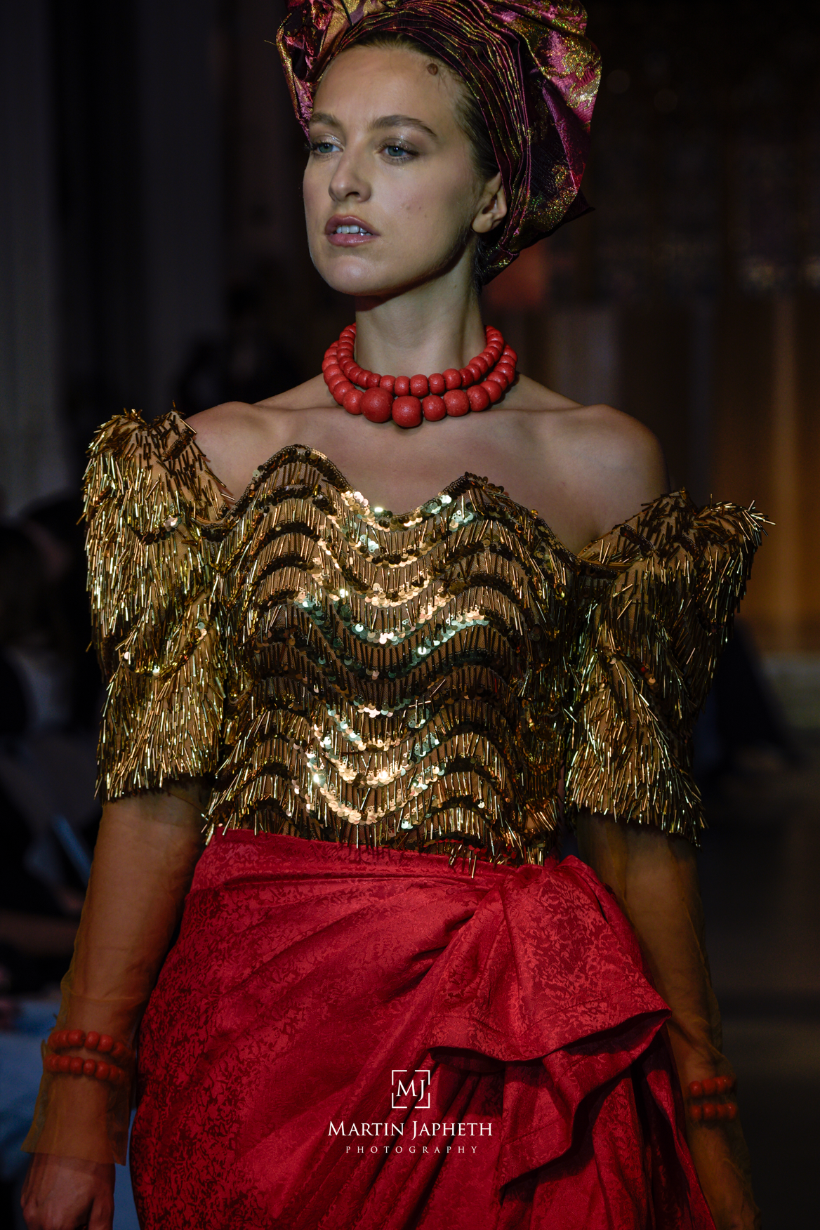Model walks runway in gold sequined top, red skirt, coral necklace, and colorful head wrap at fashion show.