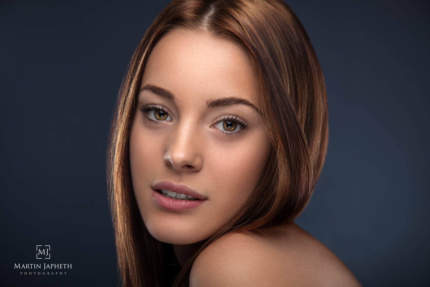 Close-up studio portrait of a young woman with brown hair, natural makeup, and a soft gaze against a dark background.
