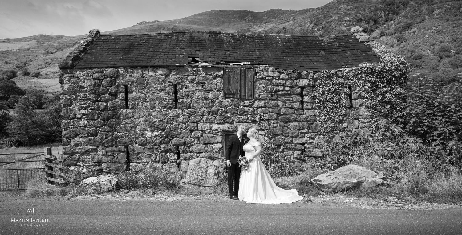 Black and white wedding photo of bride and groom embracing beside a rustic stone building in a scenic countryside landscape.