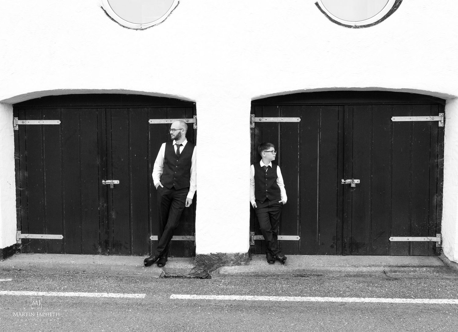 Black and white photo of two boys in formal suits posing by dark wooden garage doors on a quiet street.
