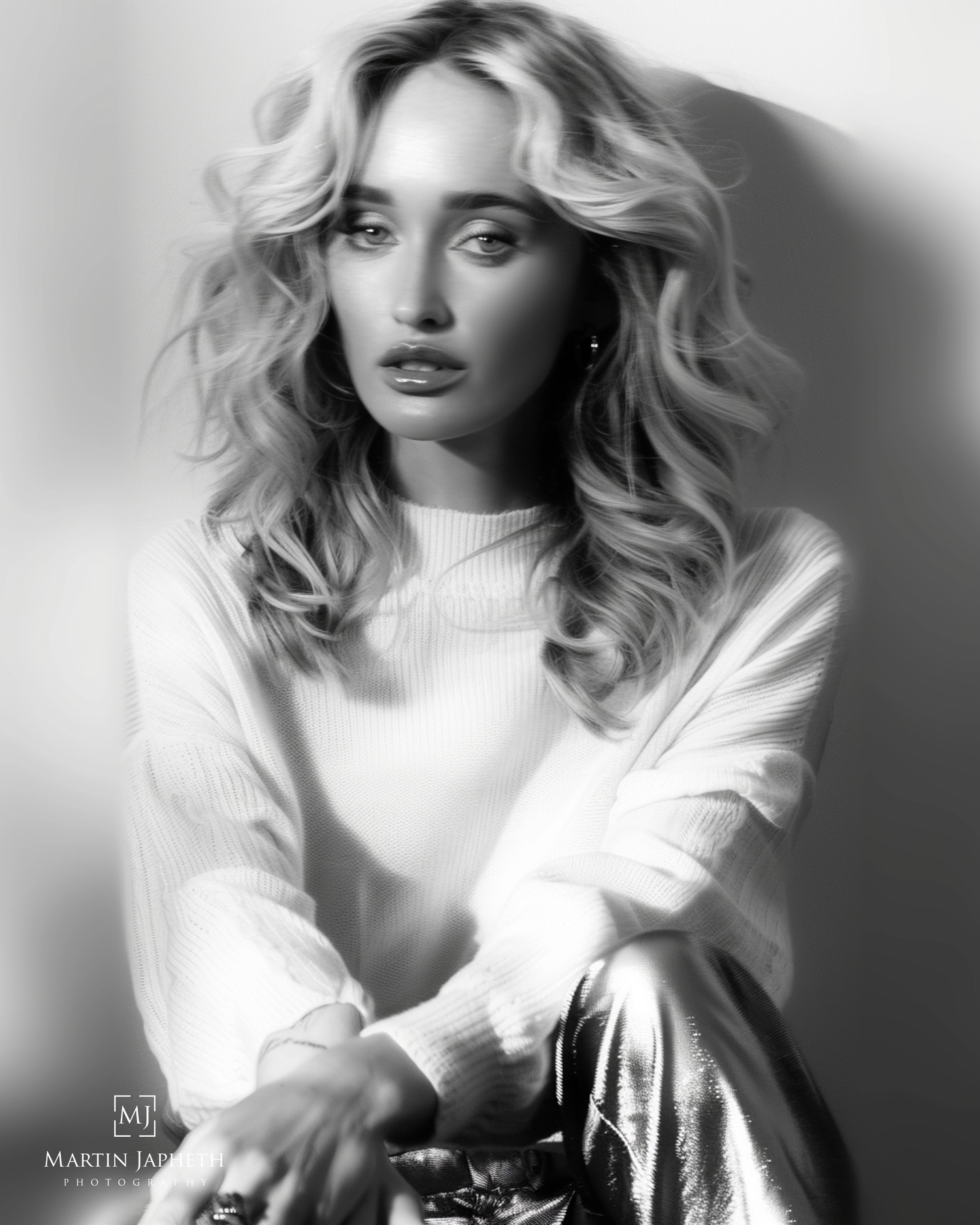Black and white portrait of a blonde woman in a white sweater and metallic pants, posing elegantly.