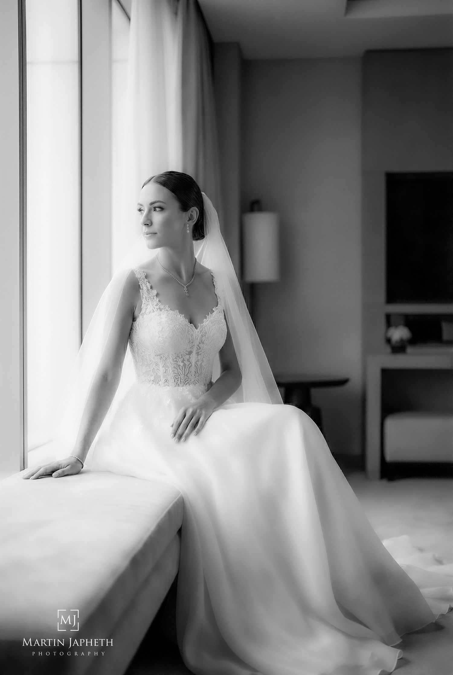 Bride in lace wedding gown and veil sits by window in black and white portrait photo by Martin Japheth.