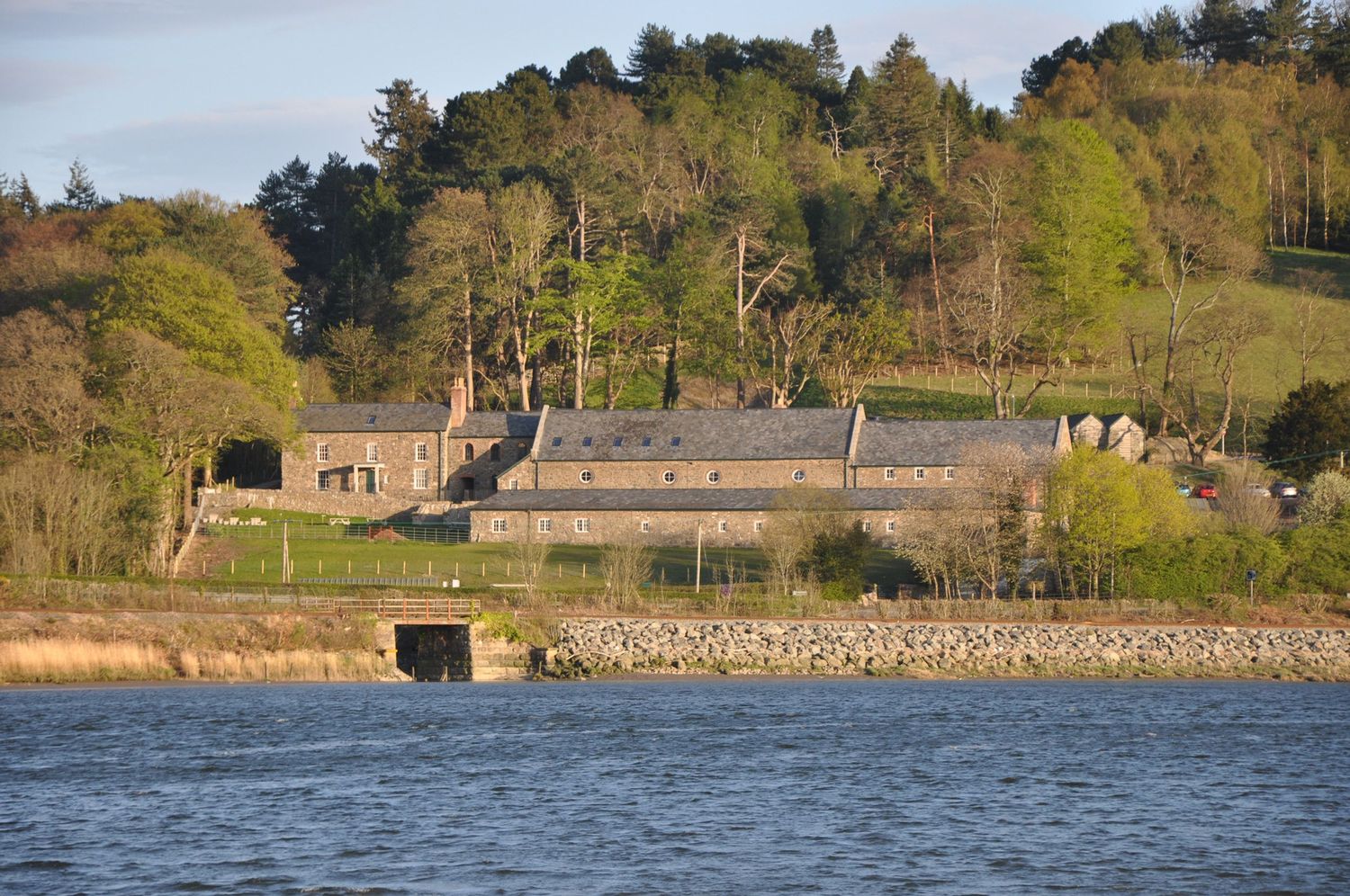 A panoramic view of a serene lake with stone buildings and trees along the shoreline in natural daylight. Hayloft at Bodnant Foods