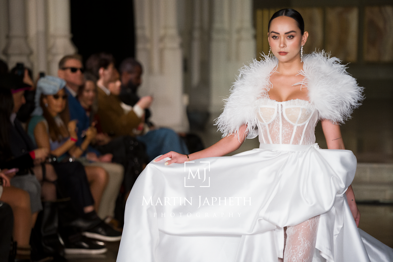 Models walk runway in white feathered bridal gowns during fashion show.