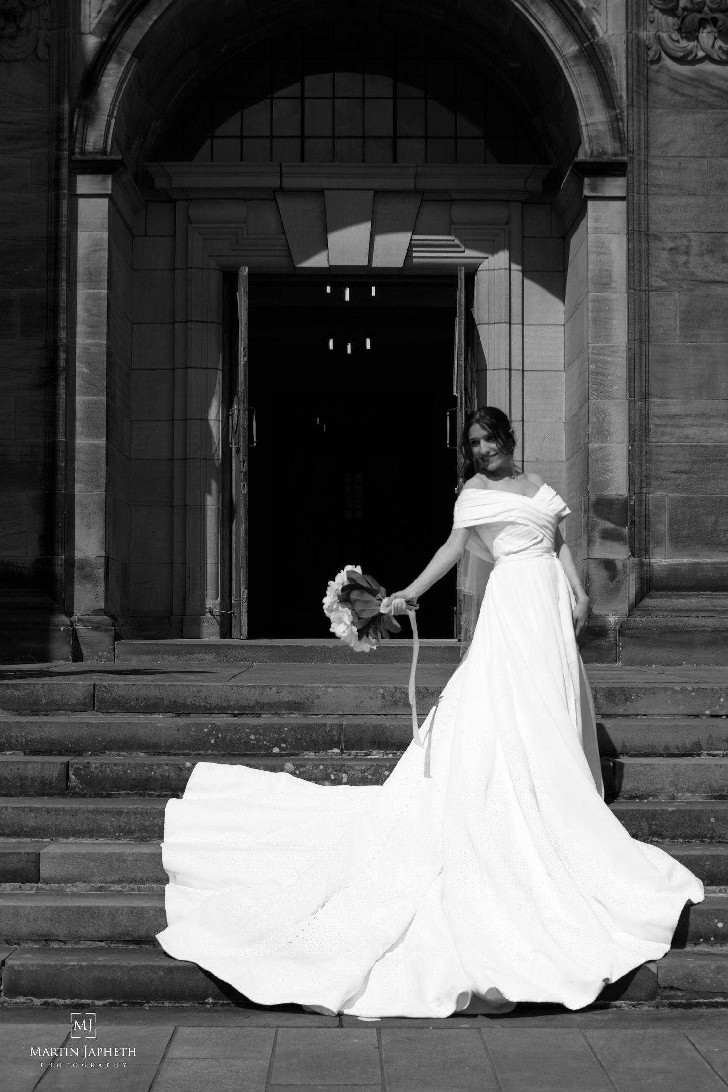 Bride in elegant white gown with train poses on church steps holding bouquet, black and white wedding photography.