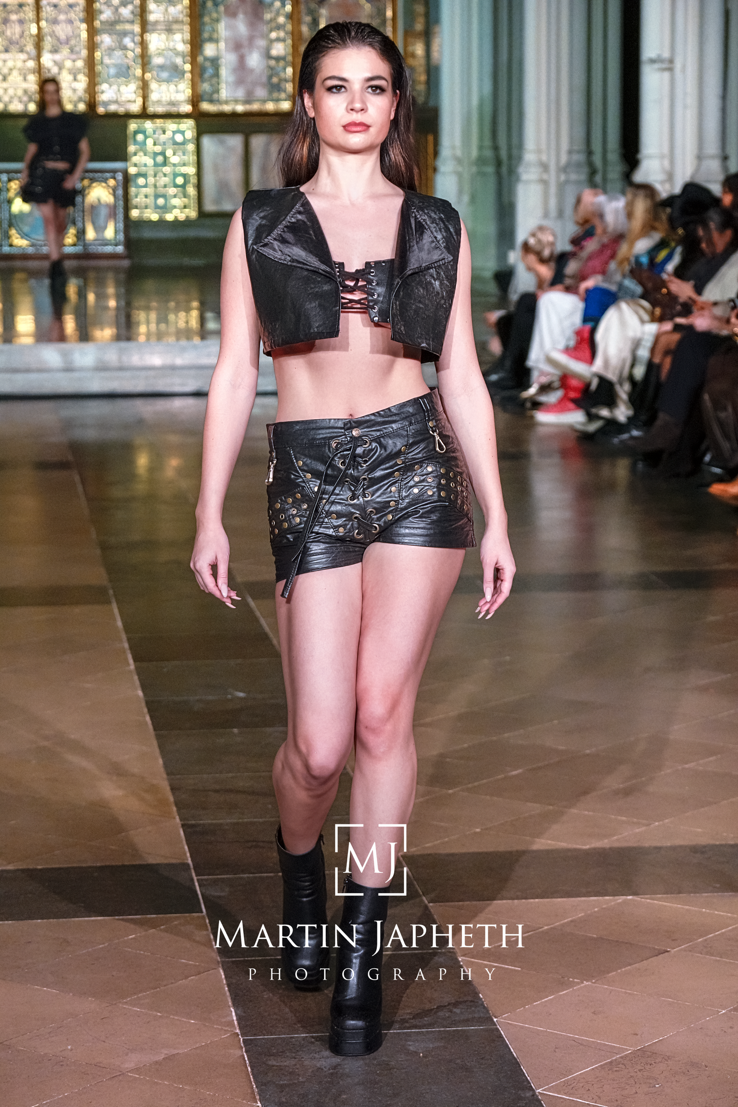 Models walking runway in black leather avant-garde fashion pieces at indoor venue with stained glass windows.