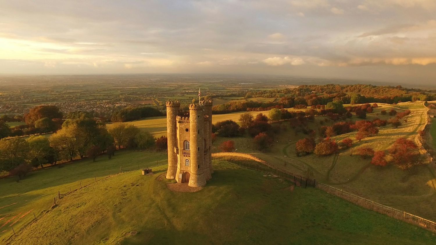 Aerial view of Broadway Tower, a historic folly atop a hill in the Cotswolds, England, at golden hour sunset.