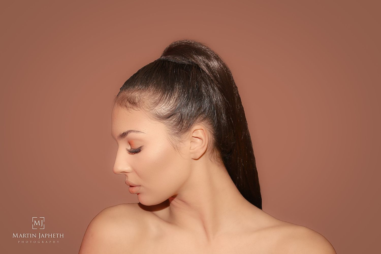 Young woman with sleek high ponytail poses in profile against warm brown background, bare shoulders, natural makeup.