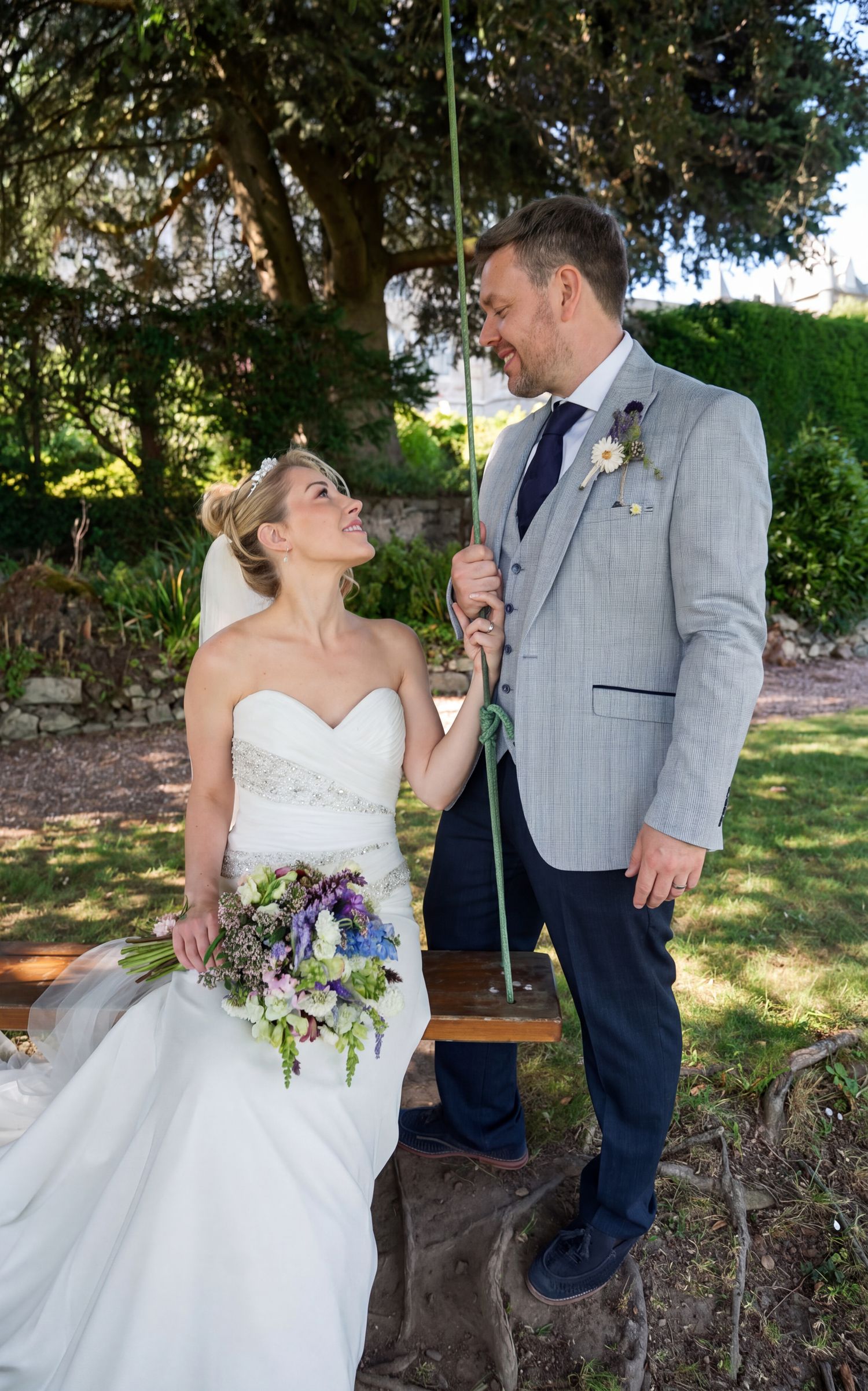 Bride sitting on garden swing holding purple bouquet gazes up at groom in grey suit at outdoor wedding.Chateau Rhianfa