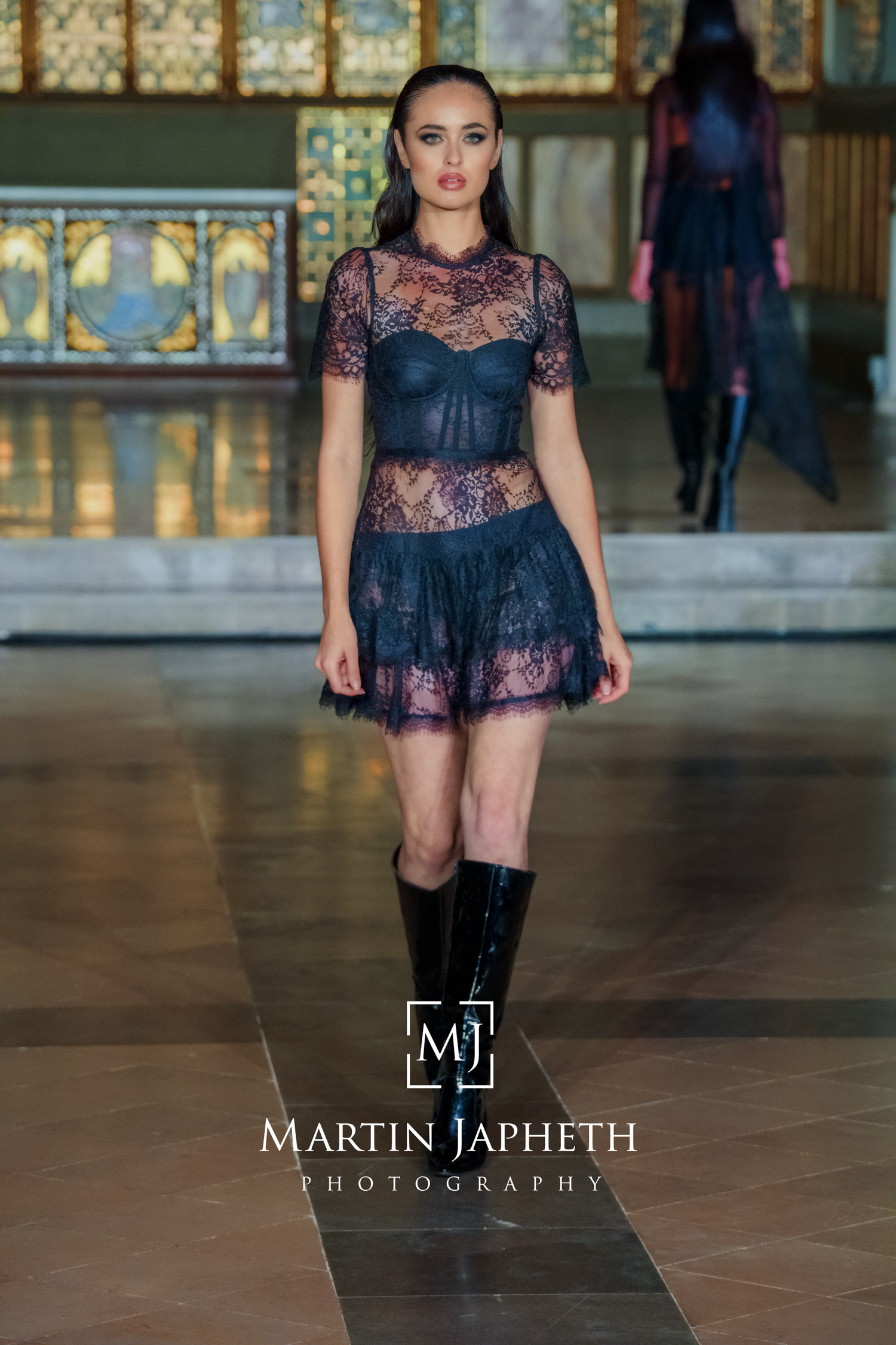 Fashion model walks runway in black lace top and knee-high boots during haute couture show.