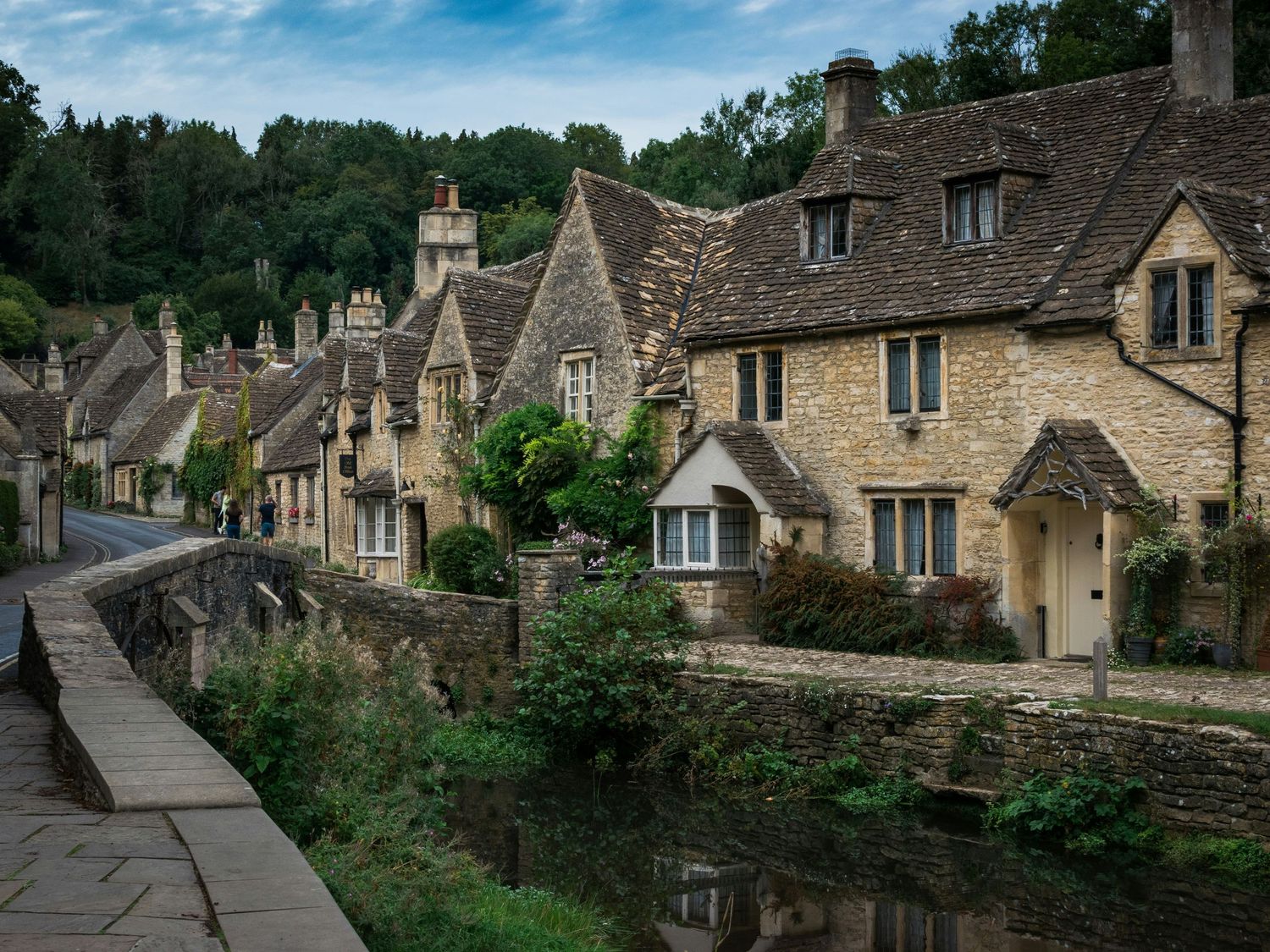 Charming Cotswolds village with honey-stone cottages lining a peaceful stream, surrounded by lush greenery in England.