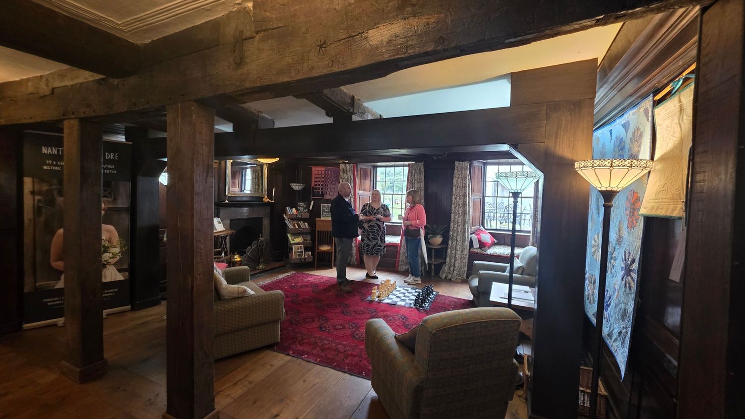 Rustic living room with dark wood beams, hardwood floors, red Persian rug, and cozy seating area with warm ambient lighting.