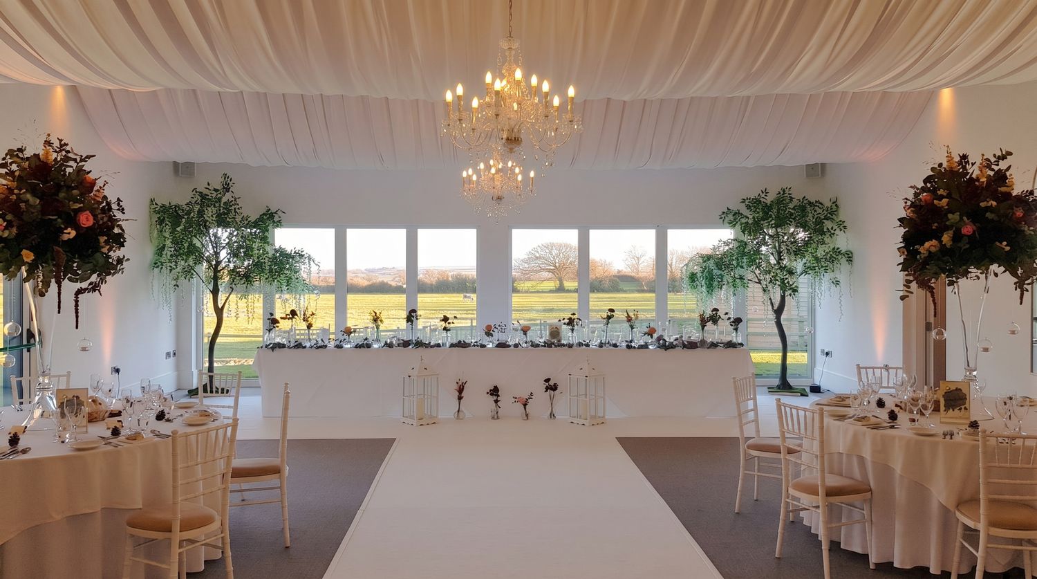 Elegant wedding reception hall with chandelier, floral trees, white aisle, and scenic countryside views through large windows.