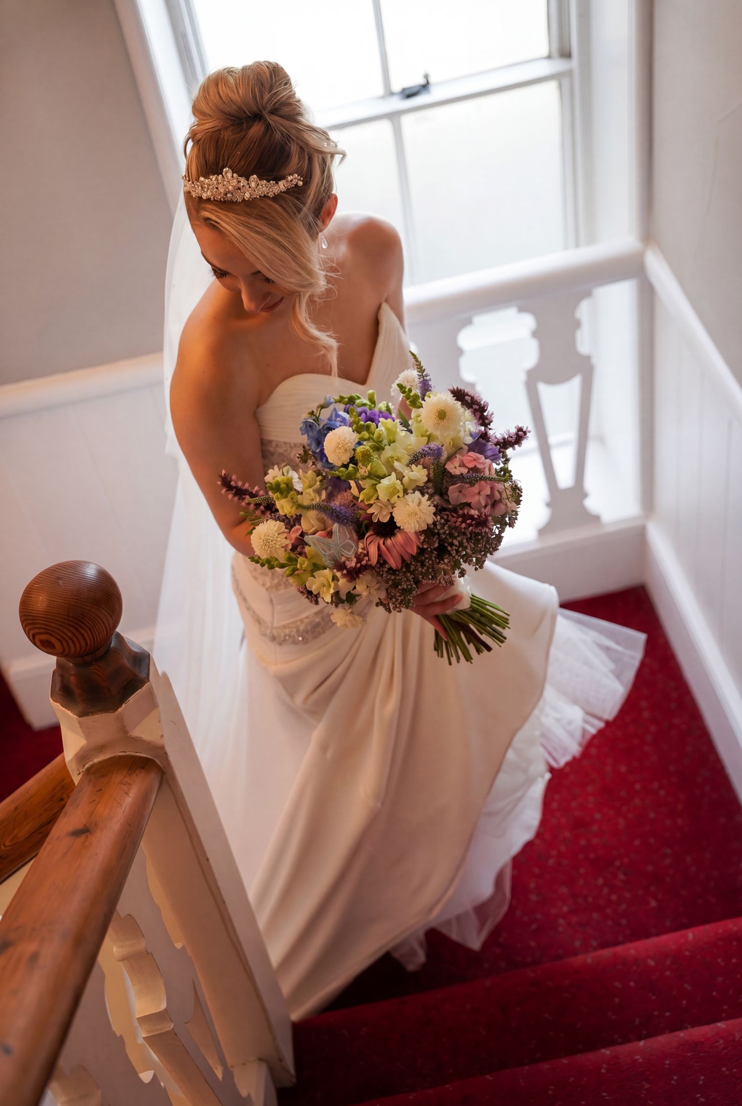 Bride in white strapless gown holding colorful bouquet descends red-carpeted staircase wearing tiara and veil at Chateau Rhianfa