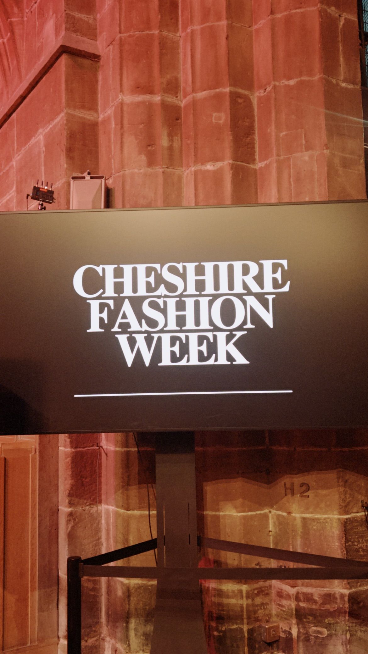 White text displaying 'Cheshire Fashion Week' against a blurred reddish-brown architectural background.