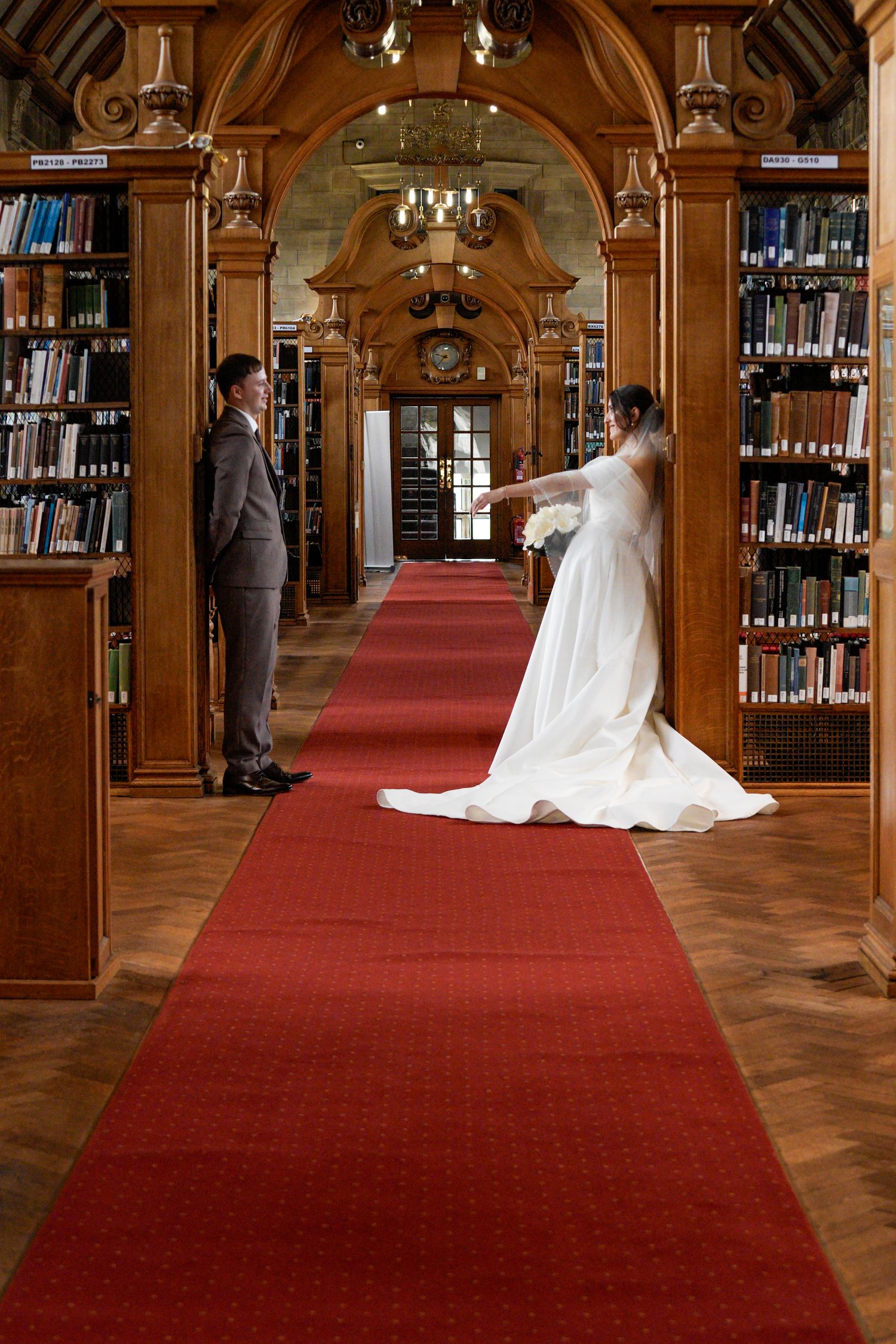 Bride and groom share a romantic first look moment in an elegant wood-paneled library with red carpet aisle at Bangor University.