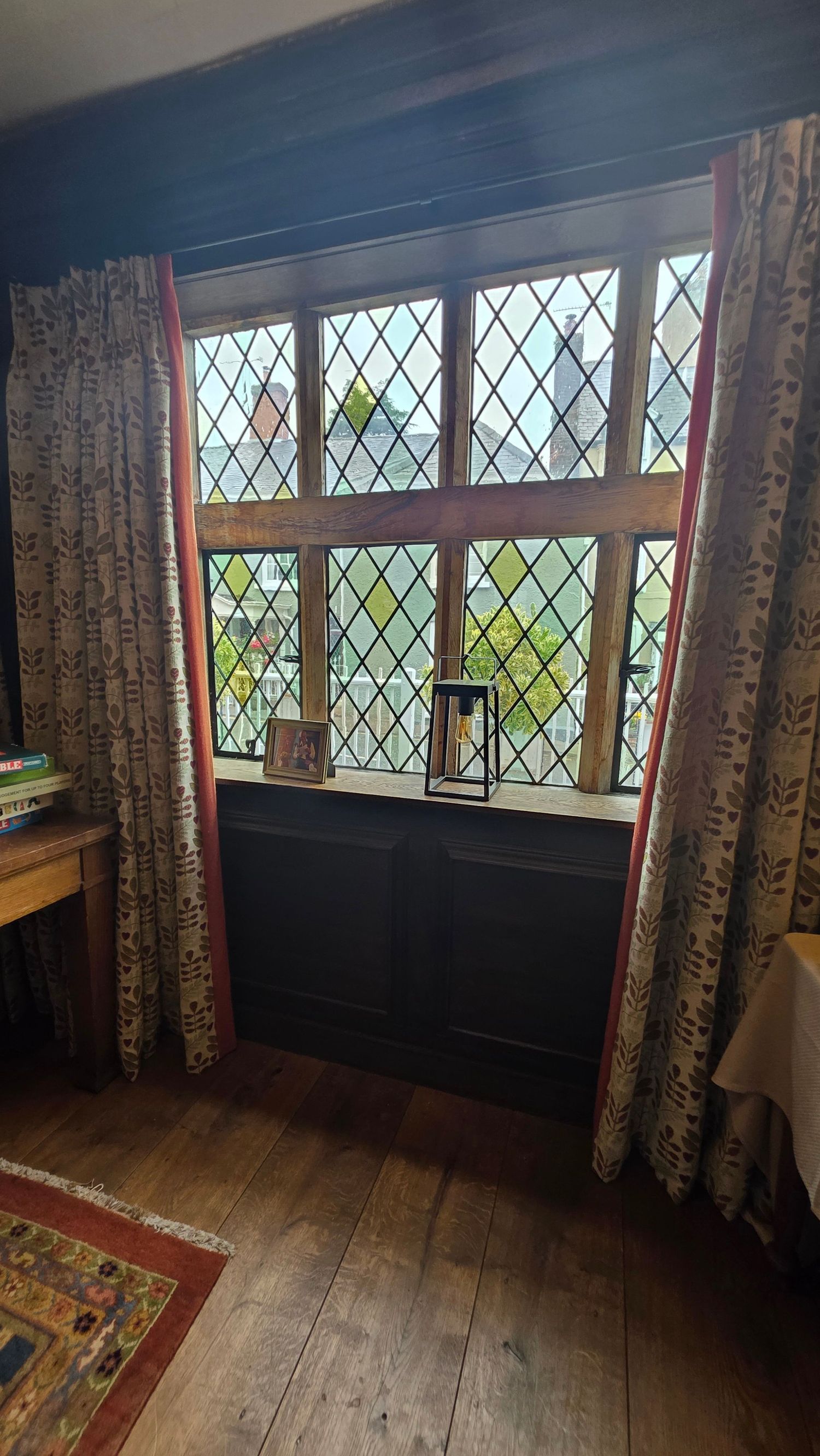 Leaded glass diamond-pane window with dark wood frame, patterned curtains, and hardwood floors in a historic interior.