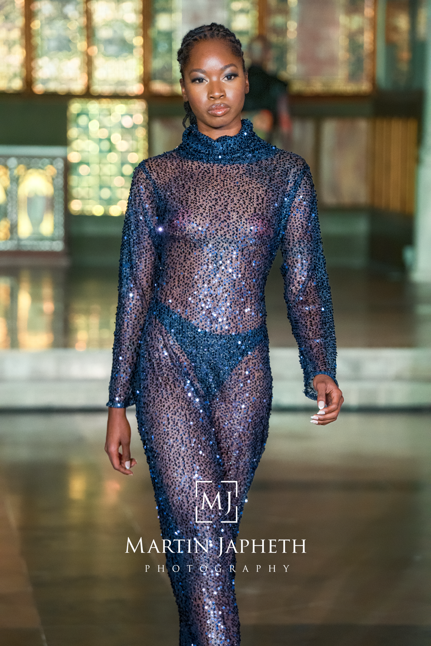 Fashion model walks runway in sheer blue sequined bodysuit with high neck during luxury fashion show.