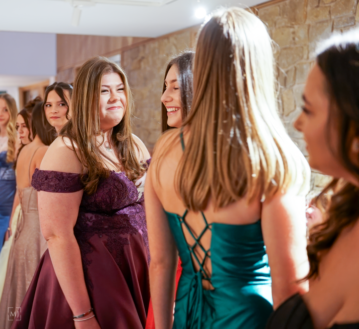 A group of women in formal dresses stand together at a social gathering, wearing burgundy and teal gowns.