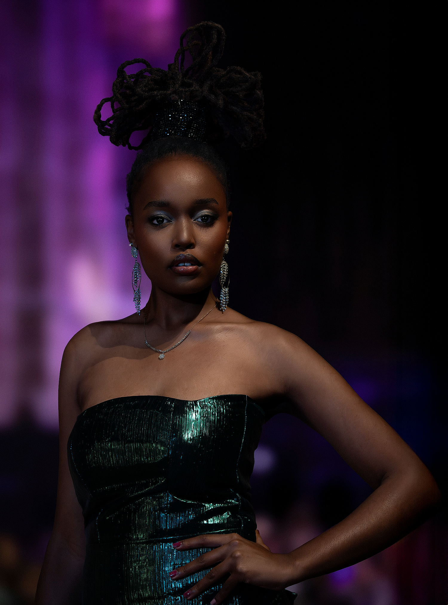 A fashion model poses in a shimmering emerald green strapless dress against a purple-lit backdrop during a runway show.