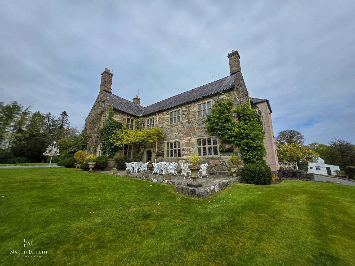 Large stone manor house with ivy-covered walls, manicured green lawn, and white garden furniture under a cloudy sky.Talhenbont Hall wedding
