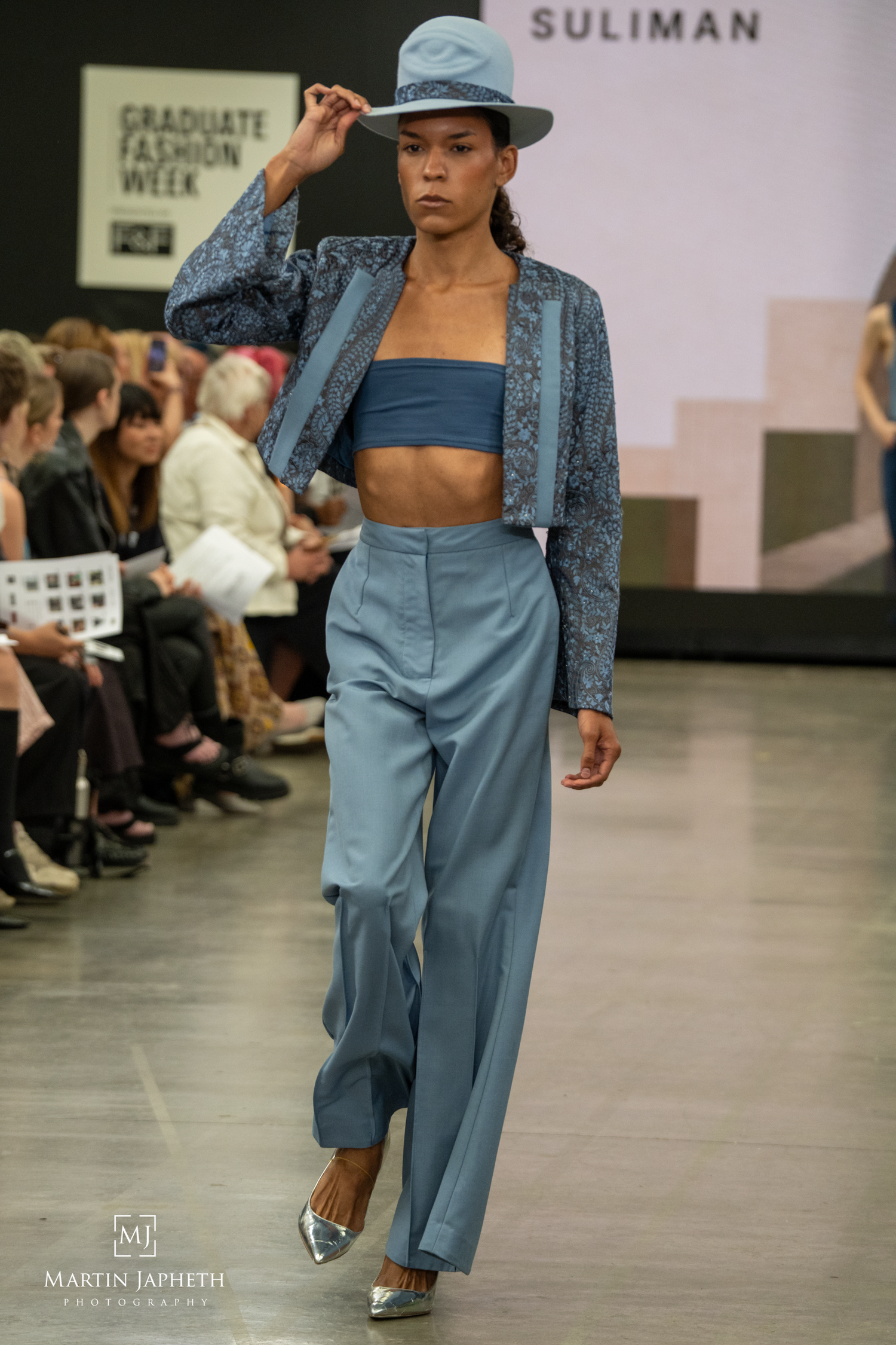 Model walks runway in blue wide-leg trousers, crop top, embroidered jacket, and hat at Graduate Fashion Week. Martin Japheth Photography
