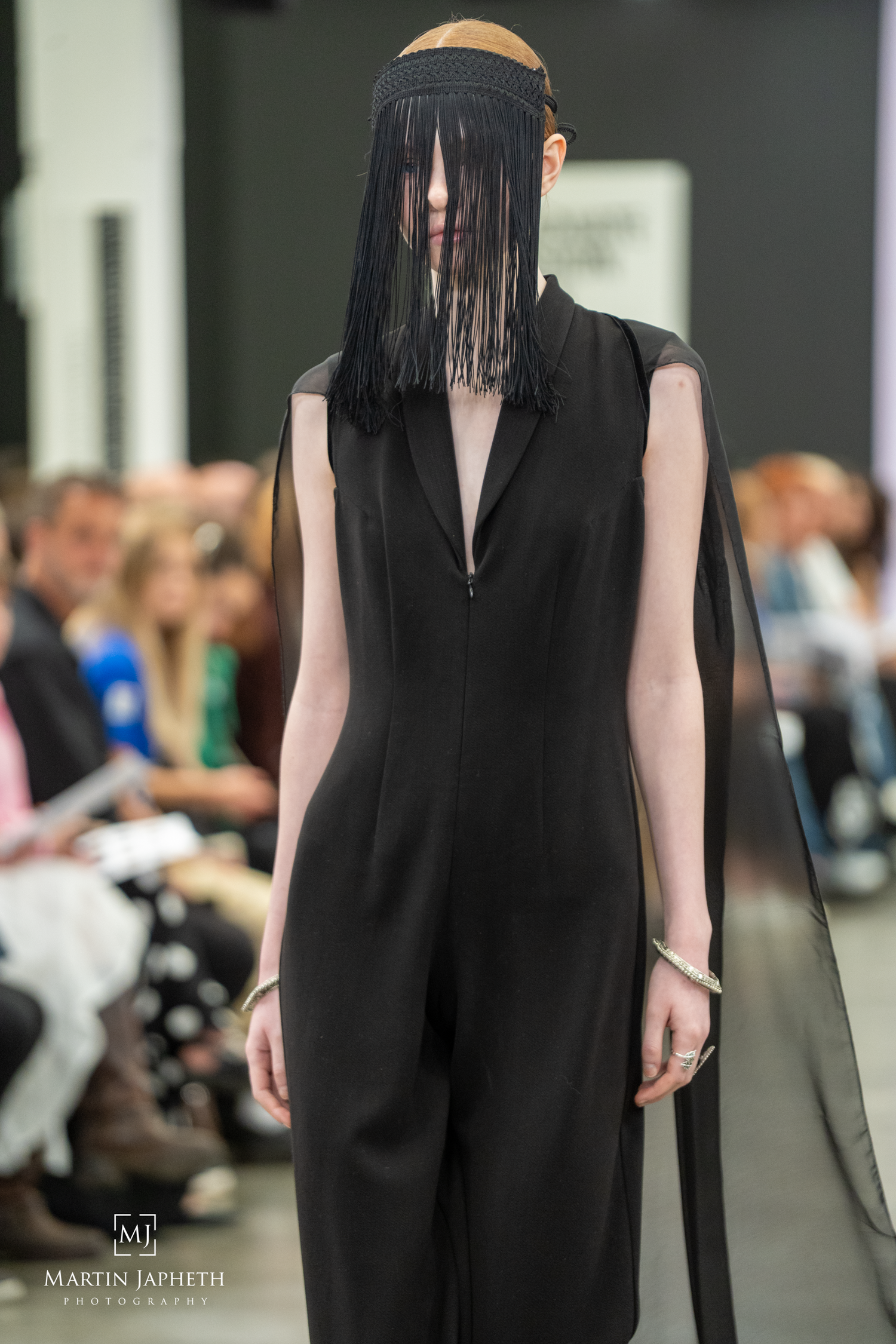 Model walks runway in black jumpsuit with dramatic fringe headpiece at fashion show. Martin Japheth Photography
