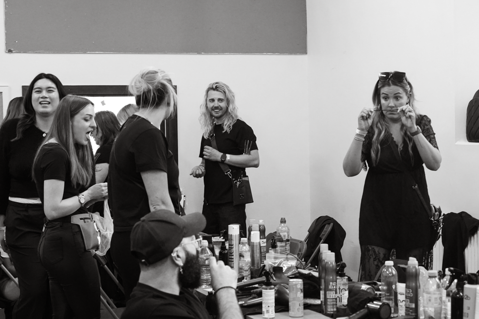 Black and white photo of stylists and models in a busy backstage dressing room with beauty products on a table.