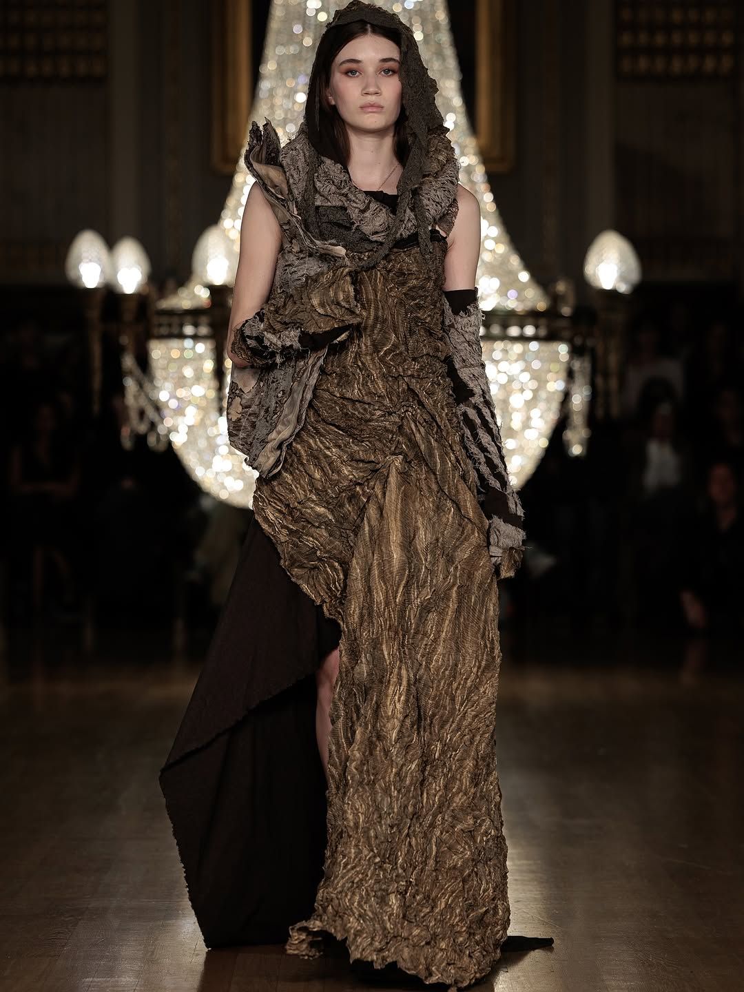 Model walks runway in textured bronze evening gown with crinkled metallic fabric during fashion show presentation.