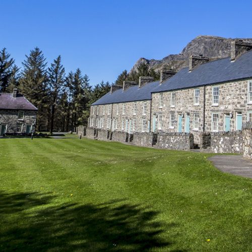 Historic stone buildings with blue doors beside lush green lawn, tall pine trees, and rocky mountain backdrop under clear blue sky.