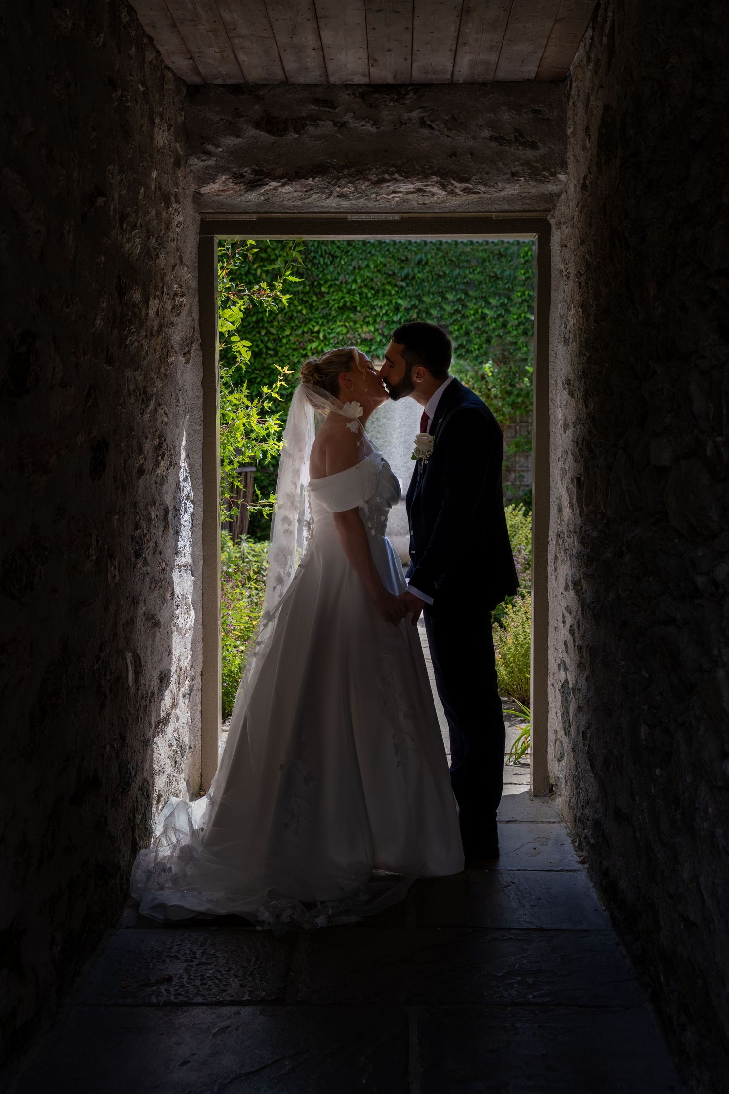Bride and groom sharing a romantic kiss in a stone archway doorway, with lush green garden visible behind them.