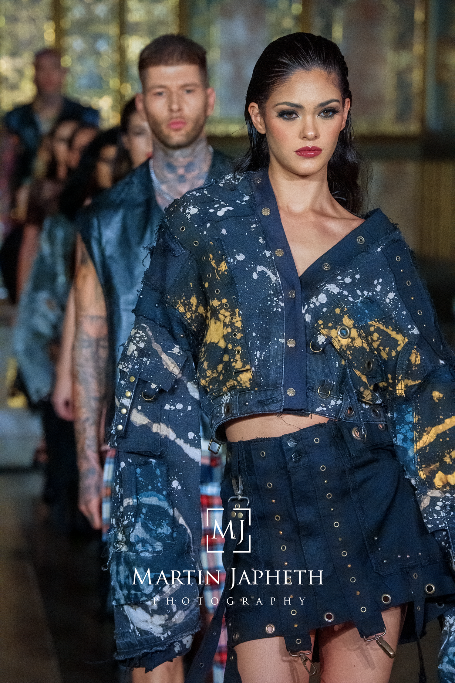 Fashion models walk runway wearing distressed denim jackets with gold paint splatter details at fashion show.