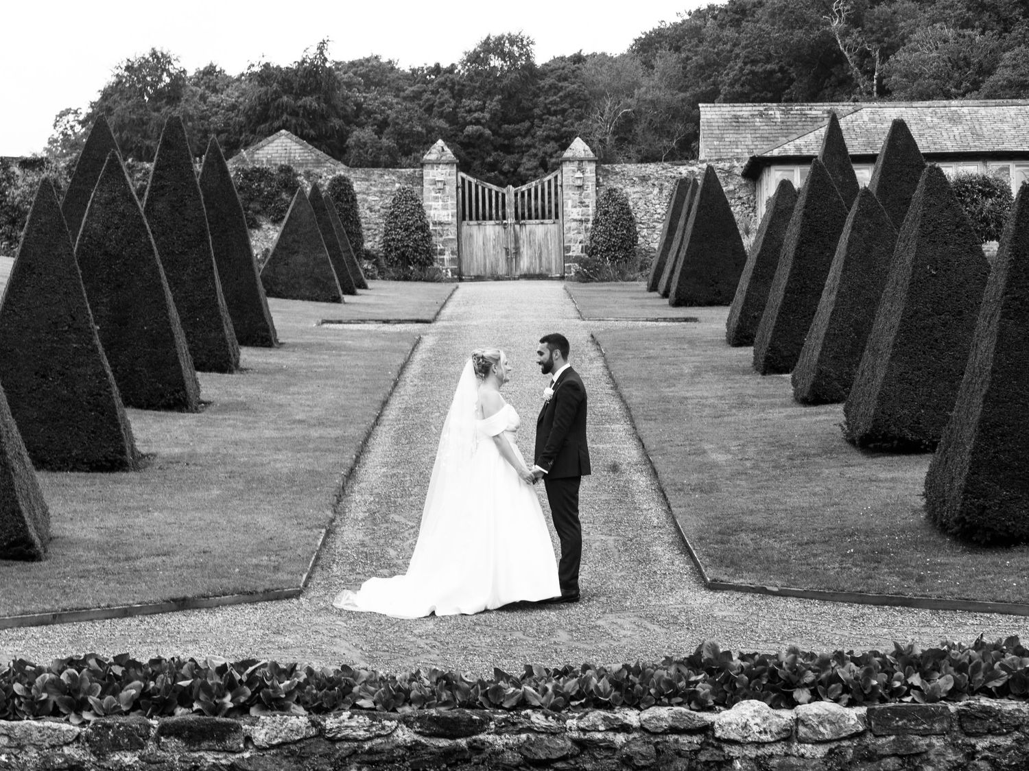 Bride in a long white gown and groom in a dark suit embrace on a garden path lined with clipped conical yew trees in black and white.