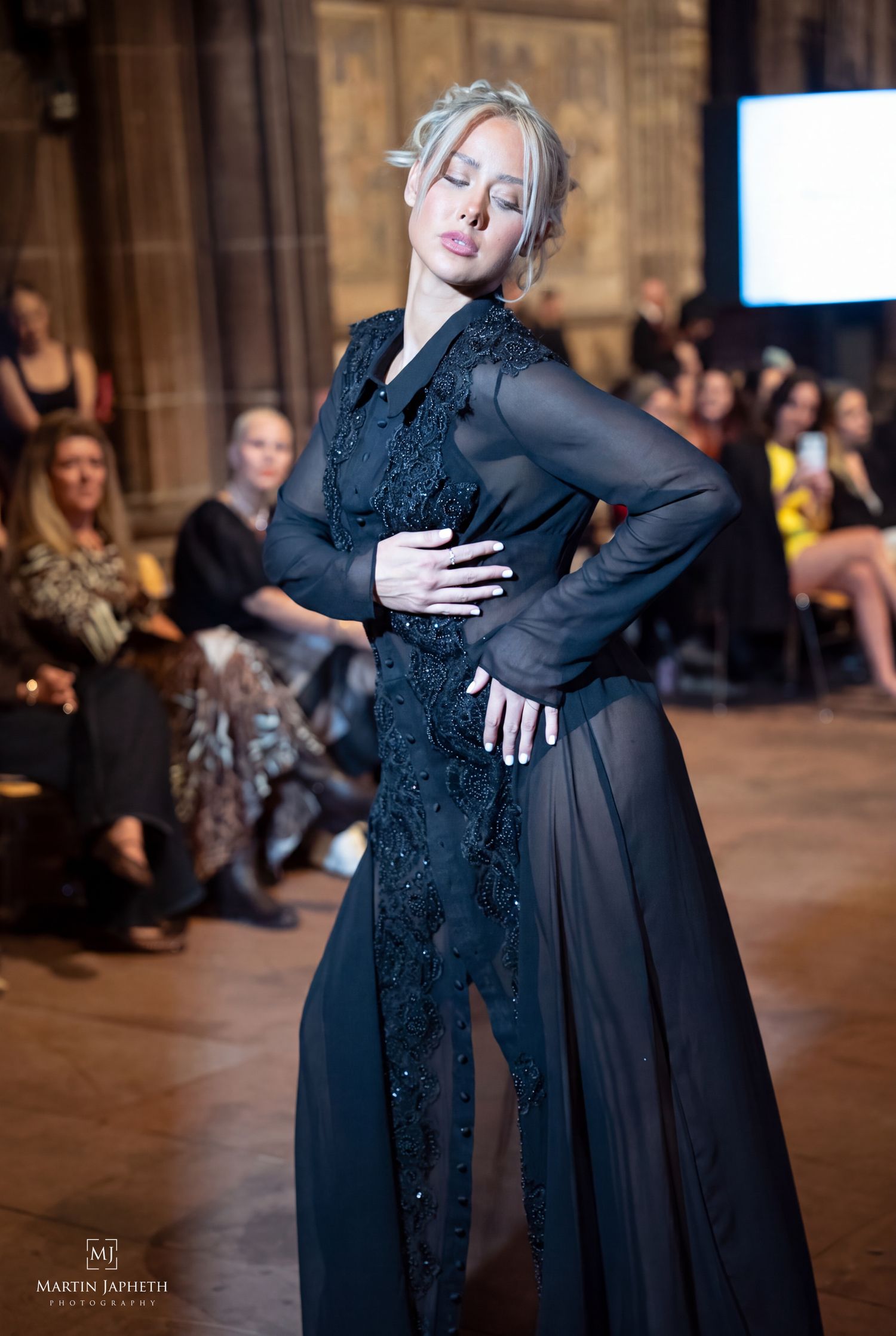 Model walks runway in elegant black sheer lace gown at fashion show event.