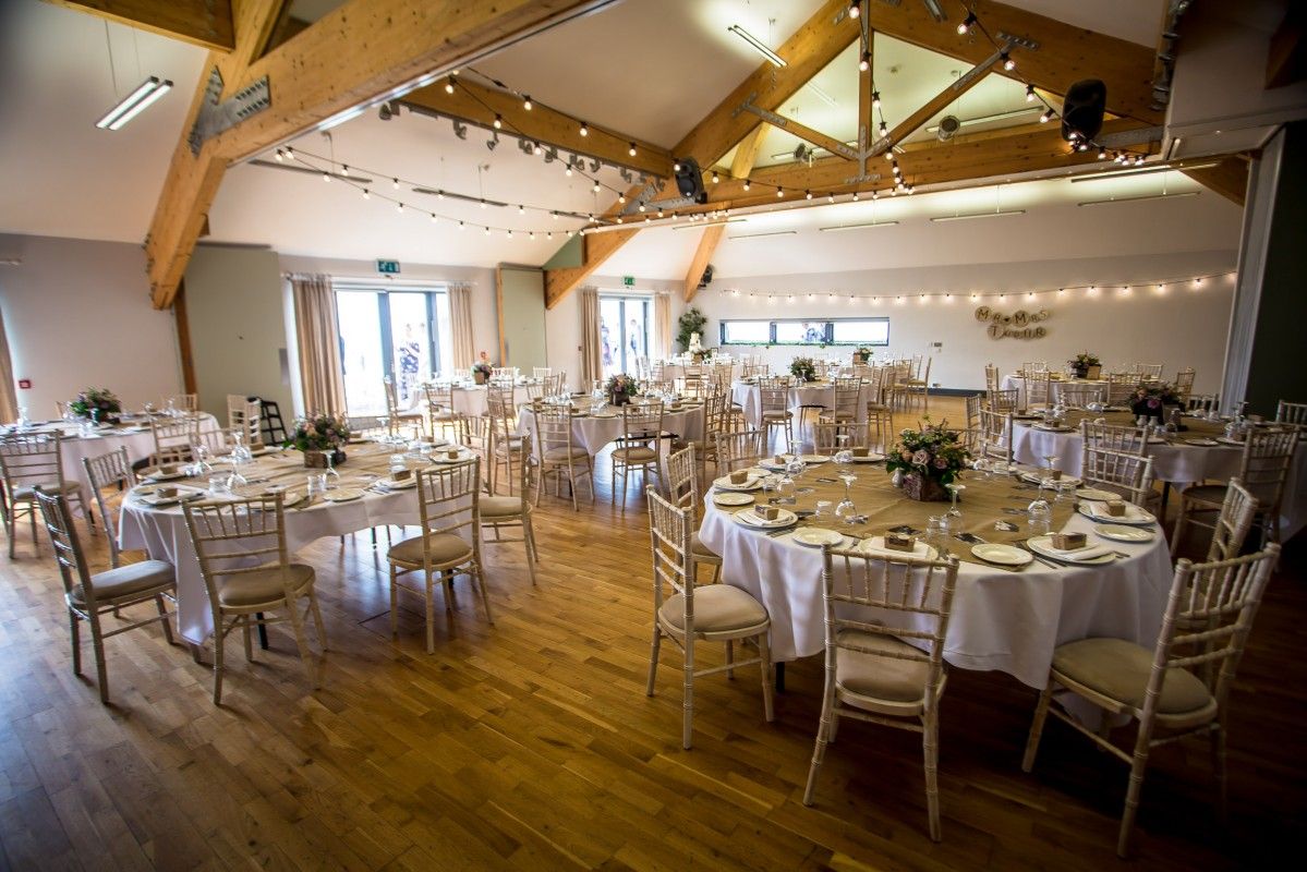 Elegant wedding reception venue with exposed wooden beams, white round tables, and string lights in rustic event space.