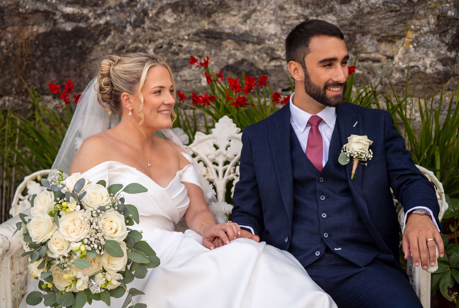 A bride and groom sit together on a white bench, she holds a bouquet, he wears a navy suit with pink tie.
