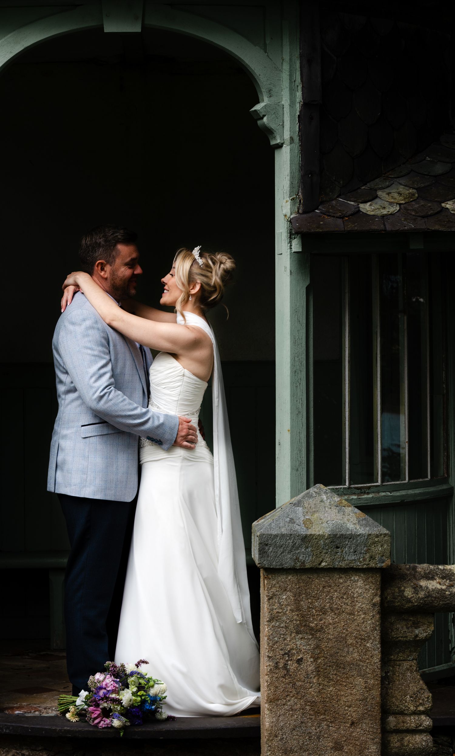 Bride and groom embracing and smiling at each other in wedding attire, standing by stone steps with bouquet nearby.