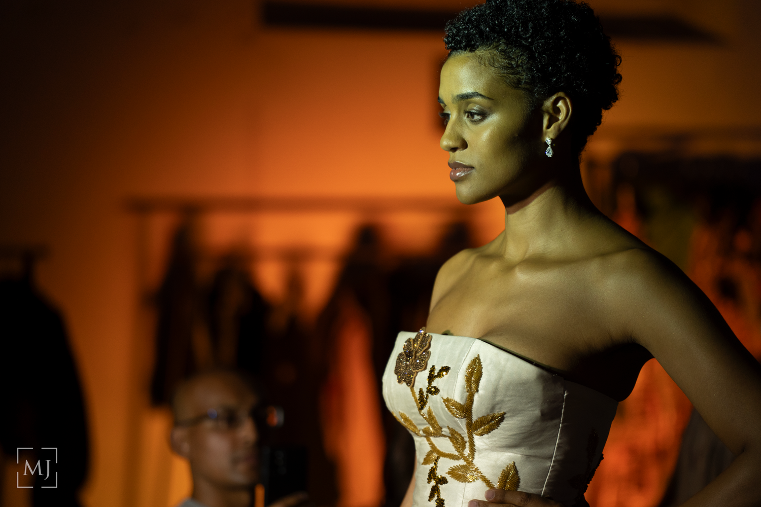 Model walks runway in strapless white corset with floral detail against warm orange lighting at fashion show.
