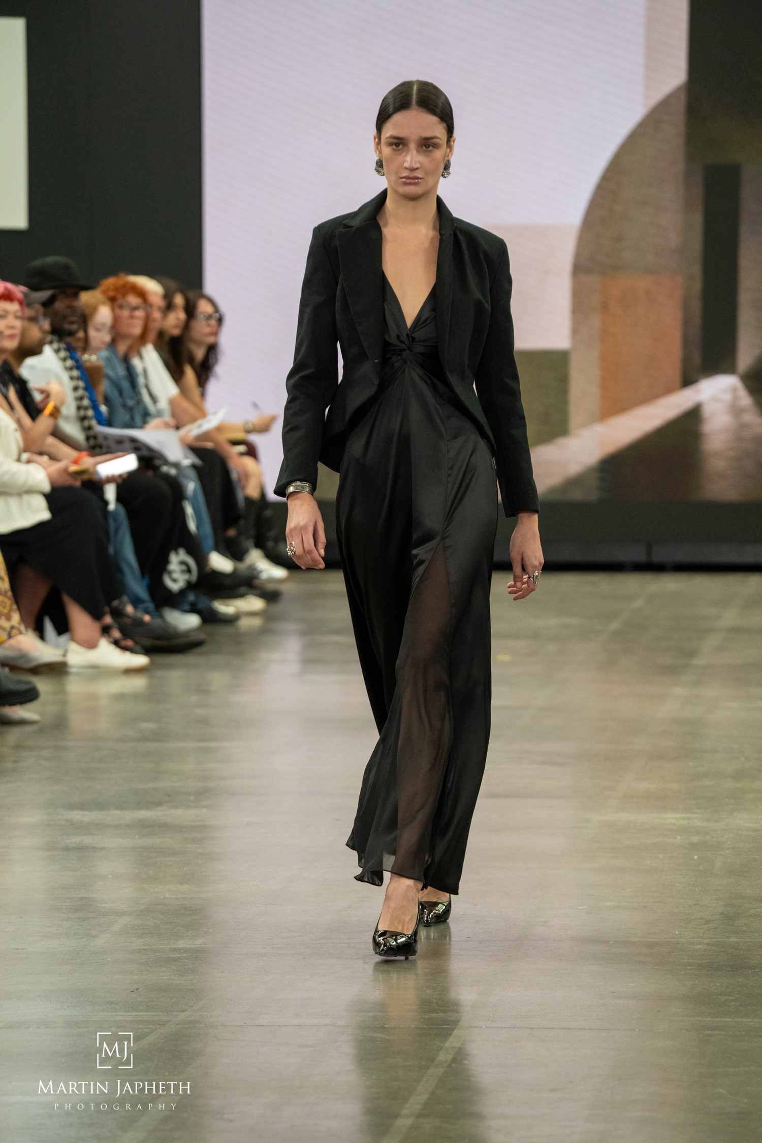 Model walks runway in black sheer maxi dress with blazer at fashion show, audience seated alongside catwalk. Martin Japheth Photography
