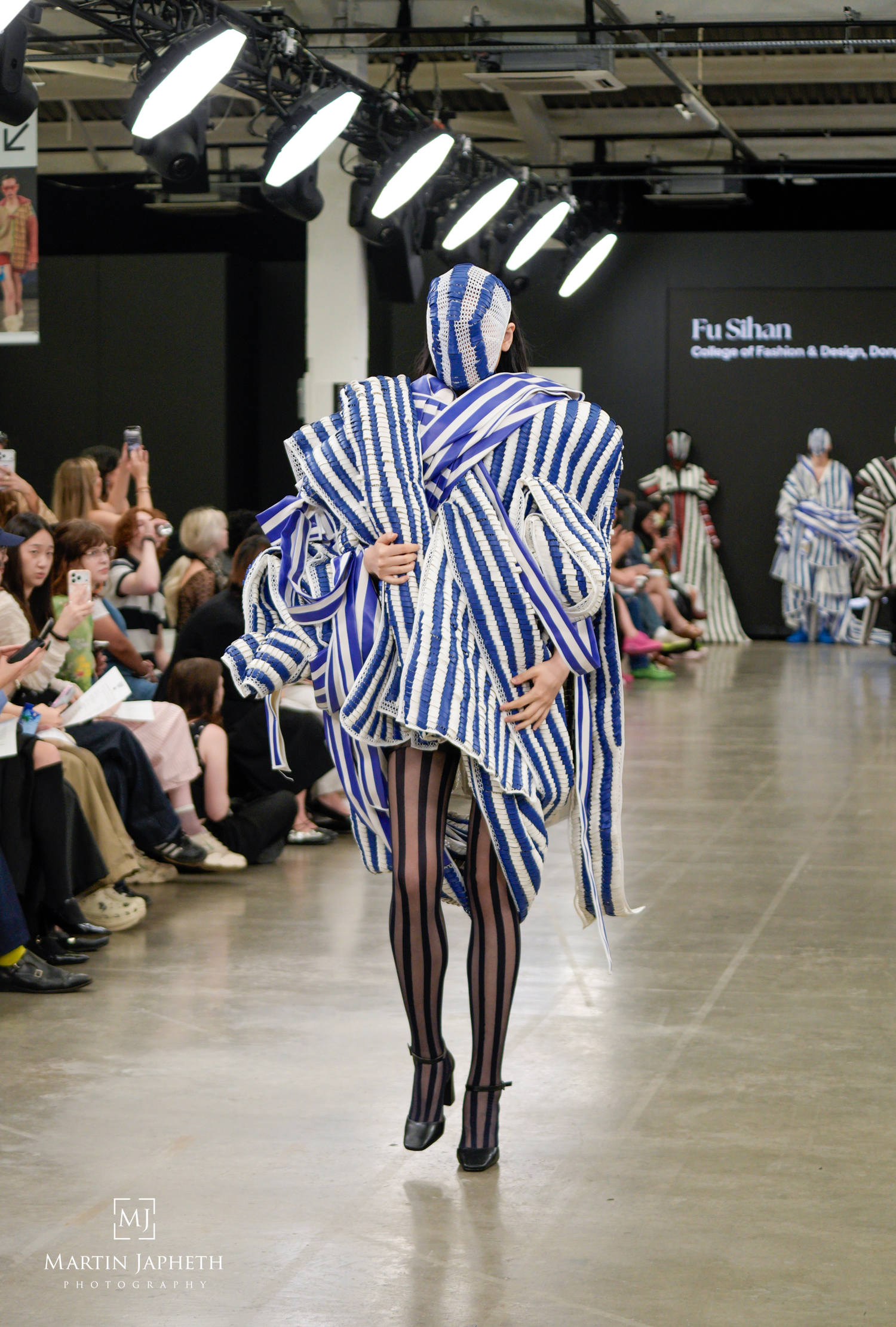 Model walks runway in bold blue and white striped avant-garde ensemble by Fu Sihan at College of Fashion and Design show. Martin Japheth