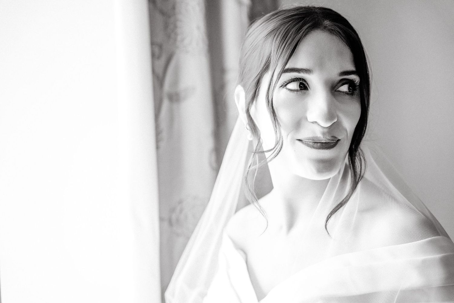 Black and white portrait of a bride in a veil gazing softly to the side near a window on her wedding day. Martin Japheth Photography