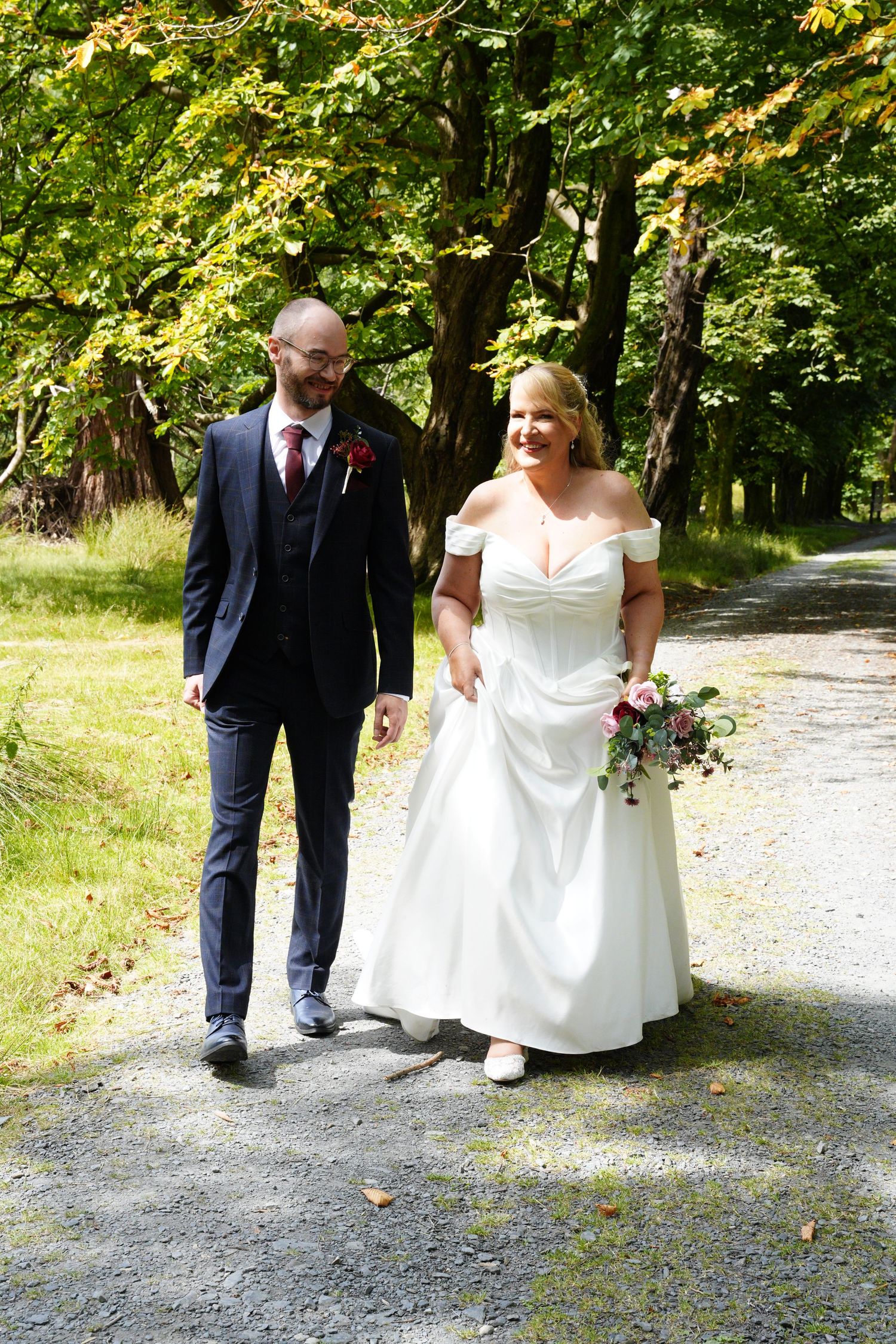 Bride and groom walking hand in hand along a tree-lined gravel path, bride in white off-shoulder gown holding pink bouquet.