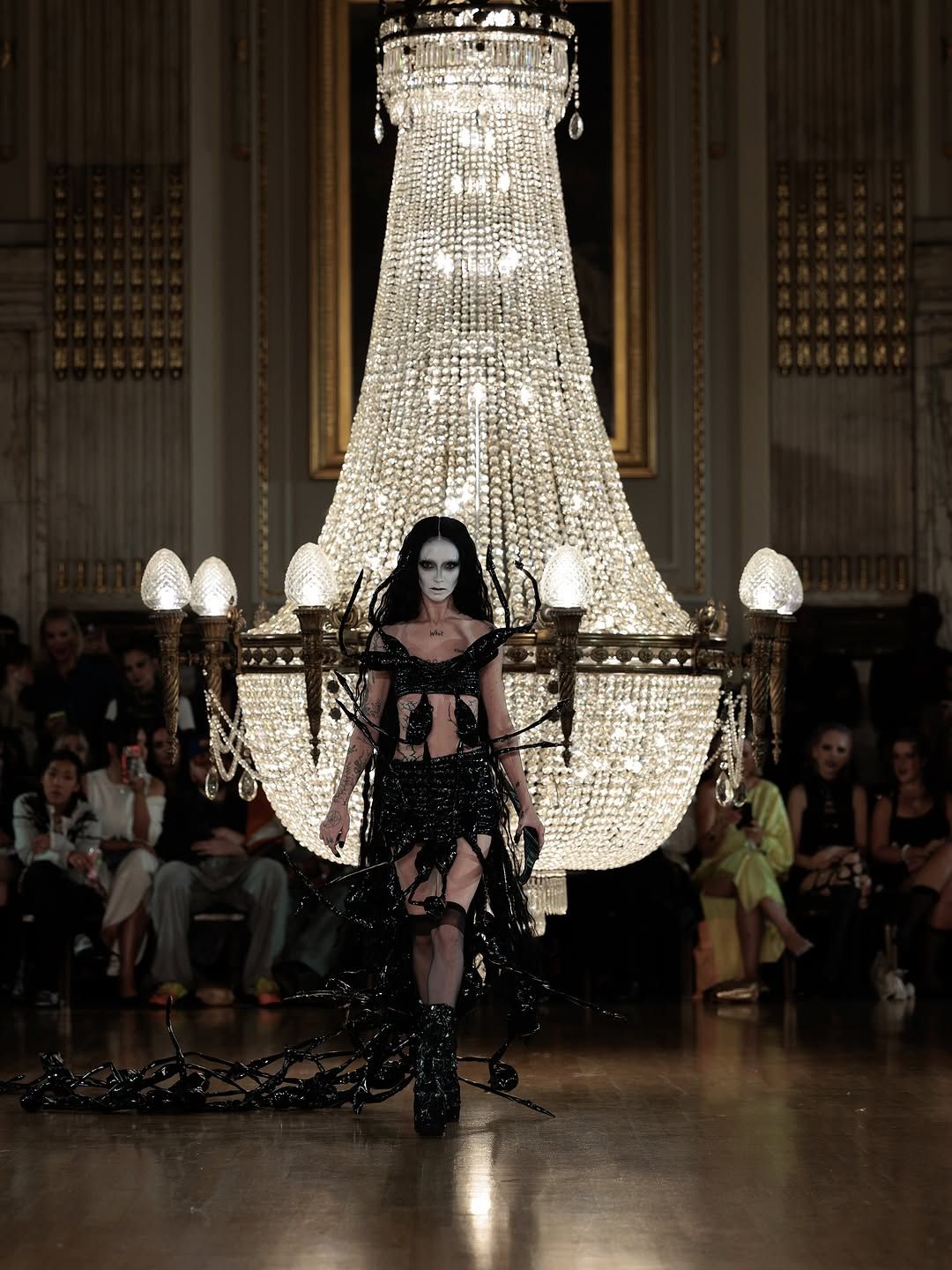 A performer hangs gracefully from an ornate crystal chandelier during a dramatic indoor performance piece.